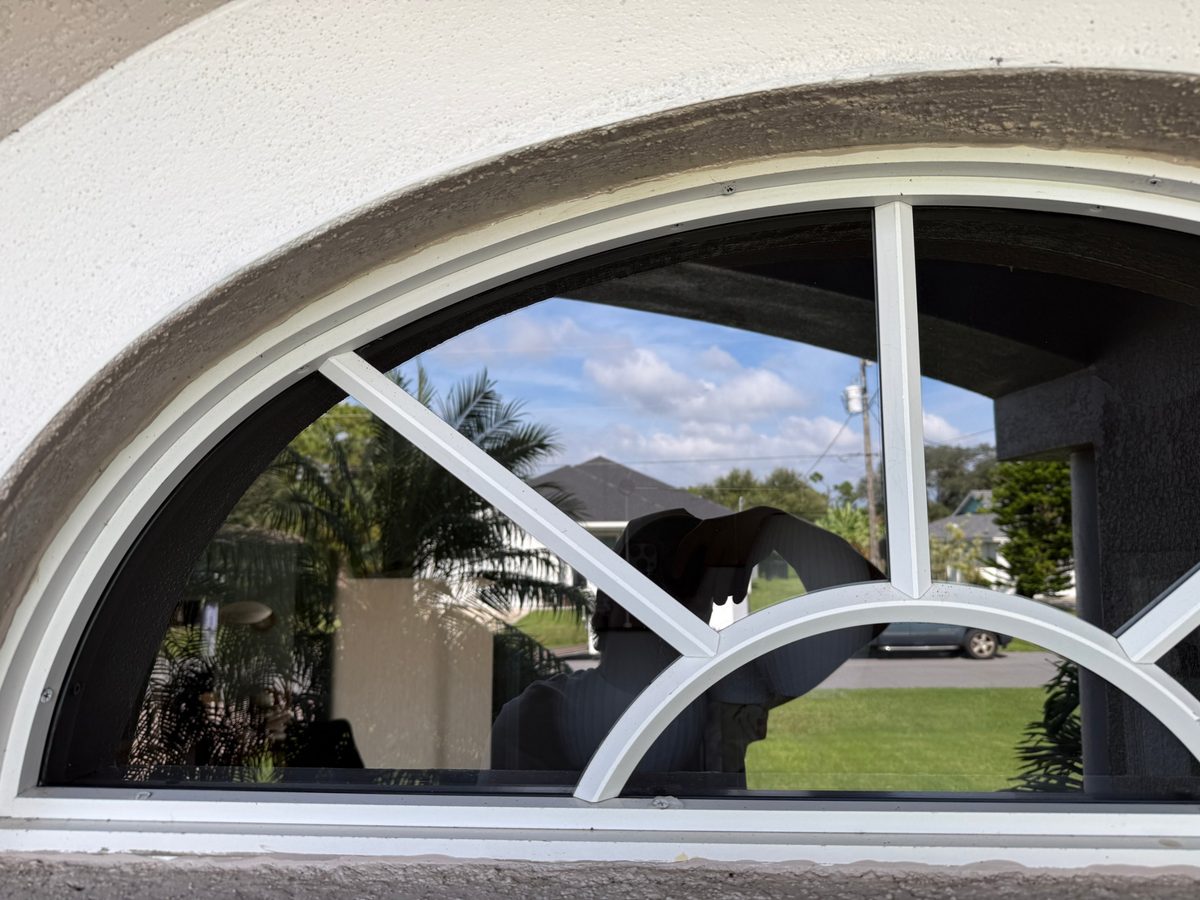 The same arched window after cleaning, with clear glass reflecting blue sky and palm fronds and a clean white frame and sill
