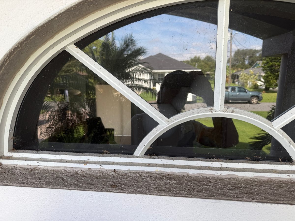 Arched fan-style window with white grille bars before cleaning, with specks on the glass and debris along the frame and sill